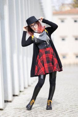 Dışarıda poz veren genç bir kadın, kırmızı ekose etekli ve şapkalı bir birey.