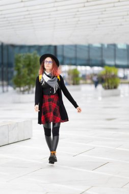 Modern şehir meydanında yürüyen gözlüklü ve şapkalı şık genç bir kadın.