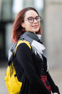 Kameraya gülümseyen, gözlük takan ve açık havada eşarp takan genç bir kadın.