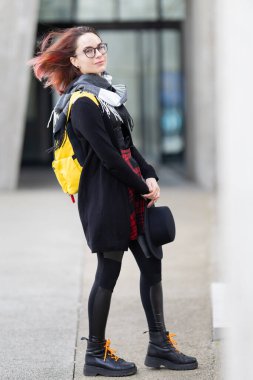 Açık havada duran, renkli saçları, sırt çantası ve modern şehir arka planına sahip şapkası olan genç bir kadın.