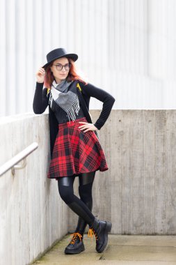 Beton duvara yaslanmış, şapka, gözlük, ekose etek ve çizme giymiş genç bir kadın.