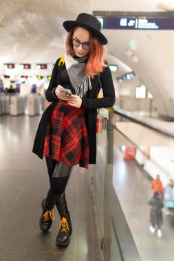 Korkuluklara yaslanan genç bir kadın. Ulaşım beklerken akıllı telefonu kontrol ediyor.