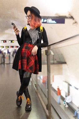 Metroda duran, akıllı telefonu kontrol eden, modaya uygun giyinen genç bir kadın.