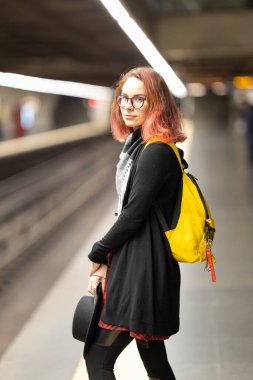 Saçları boyalı ve gözlüklü genç bir kadın metro istasyonunda toplu taşıma bekliyor.
