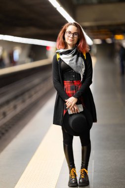 Metro platformunda elinde şapka ve telefonla sokak tarzı giyinmiş genç bir kadın.