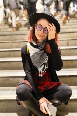 Şehir merdivenlerinde gözleri kapalı genç bir kadın elinde telefonla yorgun görünüyor.