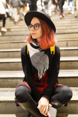 Genç bir kadın basamaklarda oturmuş, yanından geçen bulanık insanlarla düşünüyor.