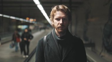 Yakışıklı, beyaz bir adam hareket halindeki bir geçitte duruyor ve kameraya gülümsüyor.