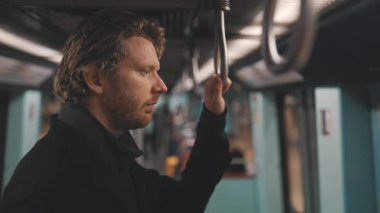 Sakallı dalgın bir adam metro vagonunda, tırabzana tutunarak seyahat ediyor.