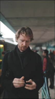 Yoğun bir tren istasyonunda bekleyen siyah paltolu genç adam.