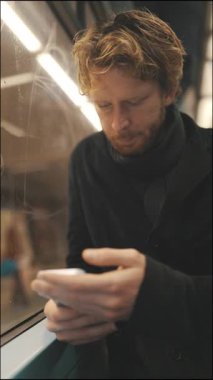 Odaklanmış sarı saçlı genç adam gece metroda seyahat ederken cep telefonunu kullanıyor.