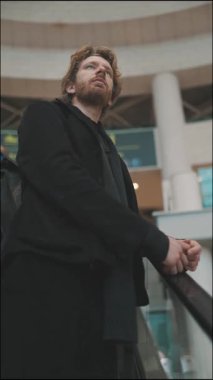 Sakallı düşünceli bir adam büyük bir kamu binasının içinde yürüyen merdivenden yukarı bakıyor.