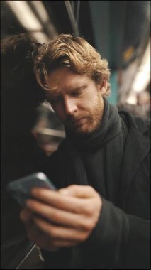 Hareket halindeki bir metroda seyahat ederken cep telefonunu kullanan sakallı sarışın bir adamın yakın çekimi.