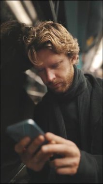 Yorgun sarışın adam metro yolculuğu sırasında pencereye yaslanırken cep telefonunu kullanıyor.