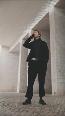 Siyah elbiseli genç adam modern beton bir köprünün altında telefonla konuşuyor.