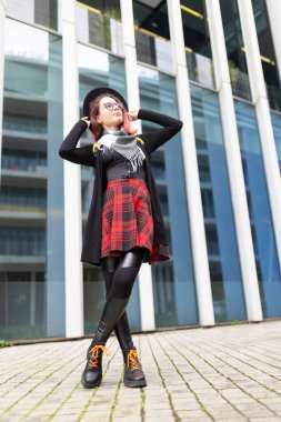 Genç kadın, ekose etek ve siyah botlarla modern mimariye karşı güven içinde poz veriyor.