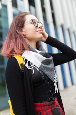 Genç bir kadın düşünceli bir gülümseme ile yukarı bakıyor, şehir ortamında modern günlük giysiler giyiyor.