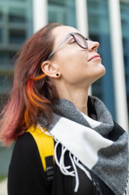 Genç bir kadın bakıyor, gözleri kapalı, rüzgarı ve güneşi şehir merkezinde hissediyor.