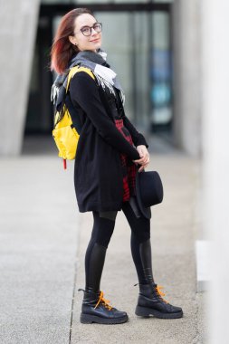 Dışarıda duran, gülümseyen, gözlüklü, eşarplı ve sarı bir sırt çantası olan genç bir kadın.