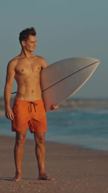 Gün batımında elinde sörf tahtasıyla kıyıda duran güzel vücutlu çekici bir sörfçü. Orta çekim