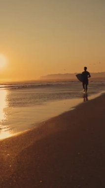 Gün batımında kıyıda koşan bir sörfçünün silueti. Orta çekim