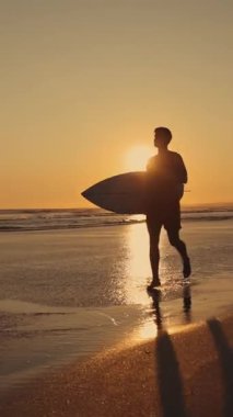 Gün batımında sahilde koşan bir sörfçünün silueti. Sonra da kameraya bakıyor. Orta çekim
