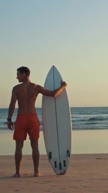 Gün batımında elinde sörf tahtasıyla kıyıda duran güzel vücutlu bir sörfçü. Orta çekim