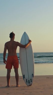 Gün batımında sörf tahtası tutan güzel vücutlu bir sörfçü. Orta çekim