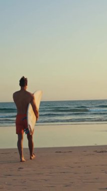 Gün batımında elinde sörf tahtasıyla okyanusa doğru yürüyen güzel vücutlu bir sörfçü. Orta çekim