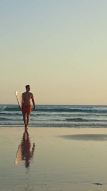 Gün batımında elinde sörf tahtasıyla okyanustan yürüyen güzel vücutlu bir sörfçü. Orta çekim