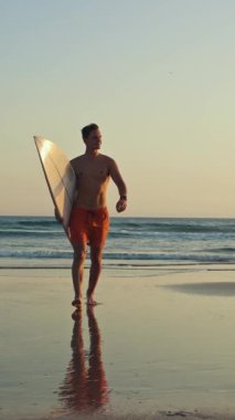 Gün batımında elinde sörf tahtasıyla okyanustan yürüyen çekici bir sörfçü kameraya bakıyor ve Shaka hareketi gösteriyor. Orta çekim