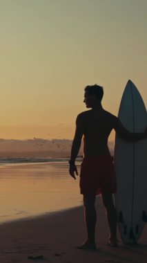 Dalgın bir sörfçü gün batımında elinde sörf tahtasıyla kıyıda duruyor. Orta çekim