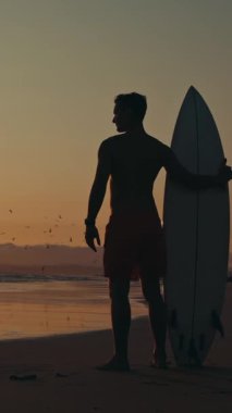 Çekici düşünceli bir sörfçü gün batımında sahilde elinde sörf tahtasıyla duruyor. Sahilde uçan martı siluetleri. Orta çekim
