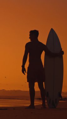 Gün batımında sörf tahtası tutan dalgın bir sörfçünün silueti. Orta çekim