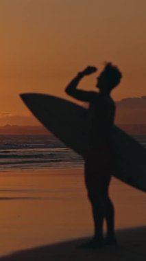 Gün batımında elinde sörf tahtasıyla kıyıda duran bir sörfçünün bulanık silueti. Orta çekim
