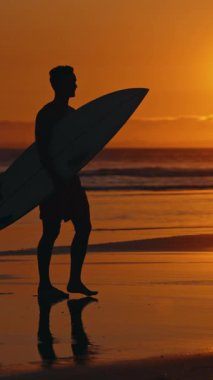 Parlak turuncu gün batımında okyanus kenarında yürüyen bir sörfçünün silüeti. Orta çekim