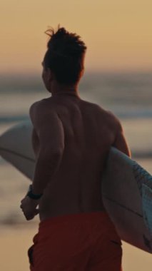 Gün batımında sörf tahtasıyla okyanus dalgalarına doğru koşan sörfçü.