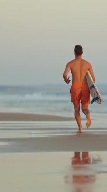 Gün batımında sörf tahtasıyla okyanus dalgalarına koşan sörfçü.