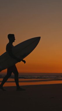 Gün batımında kolunun altında sörf tahtasıyla sahilde yürüyen genç sörfçü.