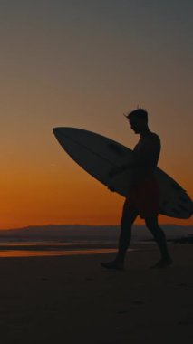 Sörfçü gün batımında sahilde yürüyor, sörf tahtası taşıyor.
