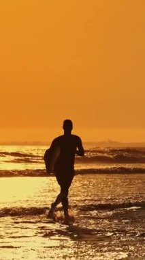 Gün batımında sörf tahtasıyla okyanus dalgalarına koşan sörfçü silueti.