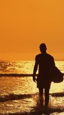 Gün batımında sörf tahtasıyla okyanustan çıkan sörfçü silueti.