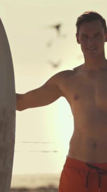 Gün batımında sörf tahtasıyla sahilde duran genç sörfçü.