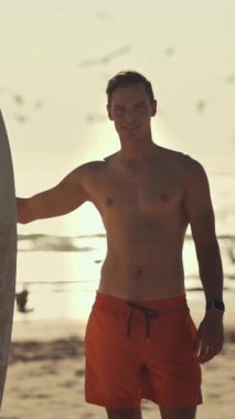 Gün batımında sörf tahtasıyla sahilde duran genç sörfçü, martılar uçuşuyor.