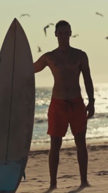 Genç sörfçü elinde sörf tahtasıyla sahilde duruyor, gün batımında uçan martıları izliyor.