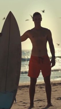 Gün batımında sörf tahtasıyla sahilde duran genç sörfçü, arka planda uçan kuşlar.