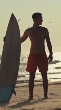 Gün batımında sörf tahtasıyla sahilde duran genç sörfçü okyanusa bakıyor.