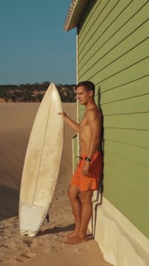Plaj kulübesinin yanında sörf tahtası tutan genç sörfçü.