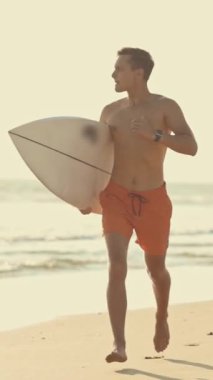 Genç sörfçü sörf tahtasıyla sahilde koşuyor.