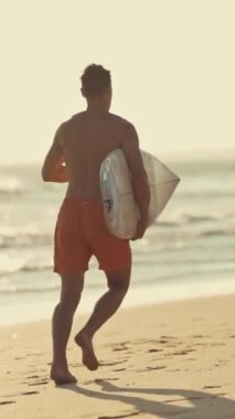 Gün doğumunda sörf tahtasıyla sahilde koşan genç sörfçü.